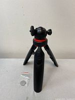 Front view of compact Mobifoto mini tripod with tripod legs deployed, black body with red accent ring on ball head assembly, paired with remote shutter and coin battery in a small plastic bag