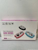 Box showing the packaging of The Penguin Dog Bowl featuring colored versions and details about dual stainless steel inserts and non-slip base