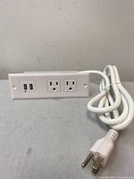 White power bar showing two USB ports and two electrical outlets horizontally aligned, with a thick white power cord and three-prong plug.