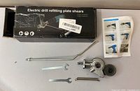 Photo of the box labeled 'Electric drill refitting plate shears' with the metal cutting attachment, wrenches, spring and instruction sheet laid out next to it.