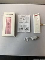 Heated eyelash curler device, USB cable, instruction manual, and product box arranged flat on a white surface.
