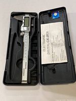Electronic digital caliper in black plastic case with instruction manual and spare battery shown in opened case.