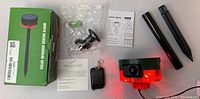 Full set photo showing the solar infrared sensor alarm with its box, mounting stakes, bracket hardware in plastic bag, remote control with keychain, and instruction sheets.