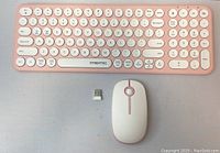 Photo showing a pink wireless keyboard with circular keys, a matching pink wireless mouse, and a USB receiver dongle on a grey surface.