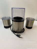 Front view of the Shardor coffee grinder with clear grinding chamber and two stainless steel containers, showing power cord and plug.