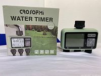 Photo showing the Crosfomi Digital Water Timer unit standing upright beside its box, clearly showing the digital display and 3 outlet connectors at the base.