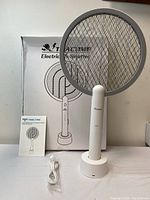 Front view of the electric fly swatter standing on its white circular charging stand. Includes USB charging cable and instruction manual to the left, box in the back.