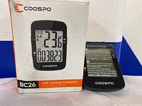 Coospo BC26 GPS cycling computer in box with device seen in front, showing the display and brand markings.
