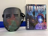 Black LED mask turned on showing colored LED smiley face pattern, next to its original retail box with a futuristic design.