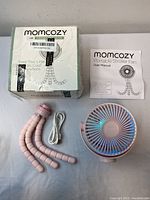Momcozy branded boxed fan, flexible segmented pink legs, USB cable, and user manual laid out on a white surface.