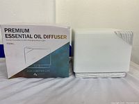 Photo showing the diffuser and its packaging box labeled 'Premium Essential Oil Diffuser' with 700ml capacity and slogan 'For a better life'. The diffuser is rectangular white and new.
