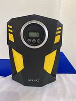 Front view of Sakaki tire inflator showing black and yellow plastic casing, central digital display with control buttons, and power button.