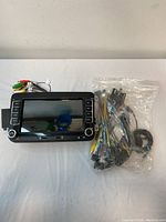 Vehicle video screen with control buttons and dual knobs, showing the front face and screen surface, accompanied by a sealed plastic bag containing wiring harnesses and connectors.