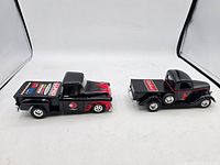 Side and rear view of two black 1:24 scale diecast bank trucks with red flame decals and sponsor logos on one and plain black with red pinstriping on the other, showing condition and details.