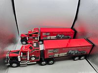 Photo showing two Buddy L semi trucks side by side with Champion Spark Plug branding on trailers, angled front view showing details of cabs and trailers.