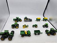 Overview of all 12 John Deere small toy tractors and farm implements displayed together on a white surface in a photo box.