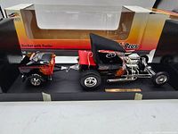 Full view of the Parts Plus 1:24 diecast T Bucket hot rod car with rear attached trailer, displayed on a black base inside original packaging box featuring yellow and red gradient with brand logo and text.