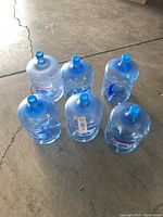 Six empty 5 gallon water bottles arranged on a concrete floor, showing their full size and handle design.