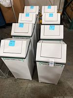 Eight white trash bins standing in two rows on a warehouse floor, tops closed, with visible blue tags on lids.