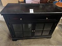 Brown wooden decor cabinet with two glass paneled doors and a drawer, viewed from front.