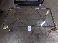 Full view of the rectangular glass top coffee table showing brass rings design and curled legs on a concrete floor.