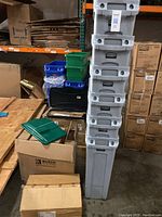 Stack of approximately 7 gray Rubbermaid Slim Jim waste baskets, showing narrow tall design in warehouse setting with other storage items nearby.