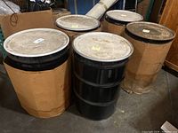 Group photo showing all five black steel drums with metal lids, one partially covered by cardboard wrap. Drums are cylindrical, approx 24" diameter x 35" height.