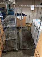 Two chrome-plated steel wire racks side by side inside a warehouse setting. Both racks have multiple wire shelves and caster wheels. Racks measure about 48 by 24 inches each.
