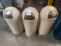 Photo showing all three beige dome-shaped trash bins with stainless steel swing-top lids placed side by side on concrete floor.