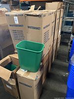 Stack of cardboard boxes labeled 'WASTEBASKET' and '12 PACK' with open boxes showing green plastic waste baskets inside