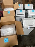 Photo of five used white swivel top trash bins standing next to two new ones packed in cardboard boxes. Plastic is visible on the new bins inside boxes labeled with MaxSold stickers.