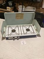 Camp stove open showing two burners, white cooking surface, light green lid lifted up, foldable side wind guards, and front control knobs.