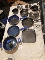 Photo showing several cookware items laid out on a white surface including frying pans with blue handles, grill pan, steamer, and saucepans.