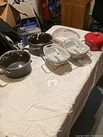 Cookware items on a table including black pots with lids, red enameled pot, white ceramic baking dishes with lids, and a clear glass baking dish.