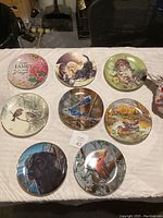 Eight decorative plates laid out on a surface, featuring animal and floral designs with gold rims.