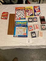Top-down view of card games, puzzle books, and cribbage board arranged on white cloth surface.