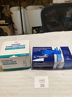 Photo showing unopened boxes of Titanline nitrile gloves and a box of hand sanitizer bottles on a table.