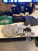 Six hoodies laid out on table, mixed sizes and colors including green Roughriders, black AC/DC, grey zip-ups and grey/black Ox Gear hoodie.