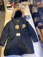 Full view of the black winter jacket with fur-trimmed hood and pom-poms, laid flat on a workbench with some tools and objects in the background.