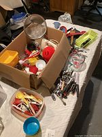 Box of assorted kitchen utensils and tools including measuring cups, peelers, strainers, scoops, and plastic containers laid out on table.