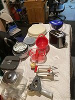 Wide angle photo showing various kitchenware items including toaster, containers, salad spinner, colander, and kitchen tools on a white cloth
