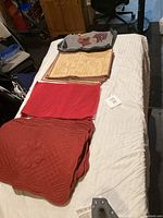 View of assorted placemats on a bed including quilted reddish placemats, red with white crosshatch, cream printed placemats and a pig-themed placemat.