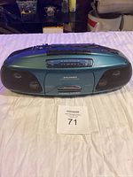 Green Sylvania portable radio with cassette player slot and front speakers shown on white cloth background with tag.