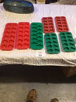 Nine silicone molds laid out on a bed, five red and four green, clearly showing the shapes of the cavities including apples and trees.