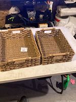 Two rectangular woven storage baskets placed side-by-side on a white table. Both have wooden handles and wicker-like woven bodies.