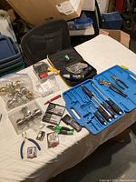 Assorted hand tools, door knobs, and hardware laid on a bed including screwdrivers, pliers, measuring tape, and packaged door knobs in clear plastic bags.