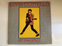 Front cover of vintage Elvis Costello vinyl record album "My Aim Is True" featuring a black and white checkered frame with a yellow center and an image of Elvis Costello playing guitar.