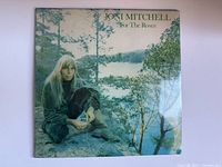 Front cover of Joni Mitchell 'For The Roses' vinyl album showing artist sitting outdoors by water and trees.