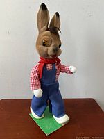 Front side of large vintage anthropomorphic rabbit figurine on green base, wearing blue overalls and red checkered shirt, white gloves, and a red scarf.