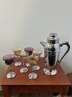 Full view of cocktail shaker with six goblets arranged on wooden table.